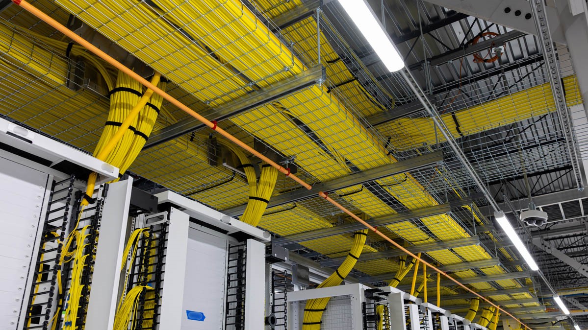 A shot looking up at the waves upon waves of yellow Ethernet cables connecting the xAI Colossus cluster to itself. Multiple layers of excessively wide cable runs are recessed into the ceiling.