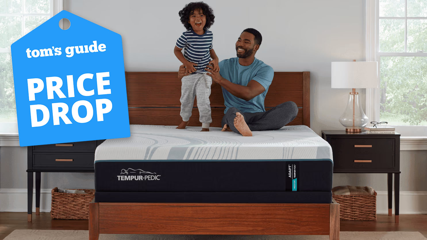 The Tempur-Pedic Tempur-Adapt mattress on a bed frame in a bedroom, a man and his young sun sitting on top of it, smiling. A Tom's Guide price drop deals graphic (right)
