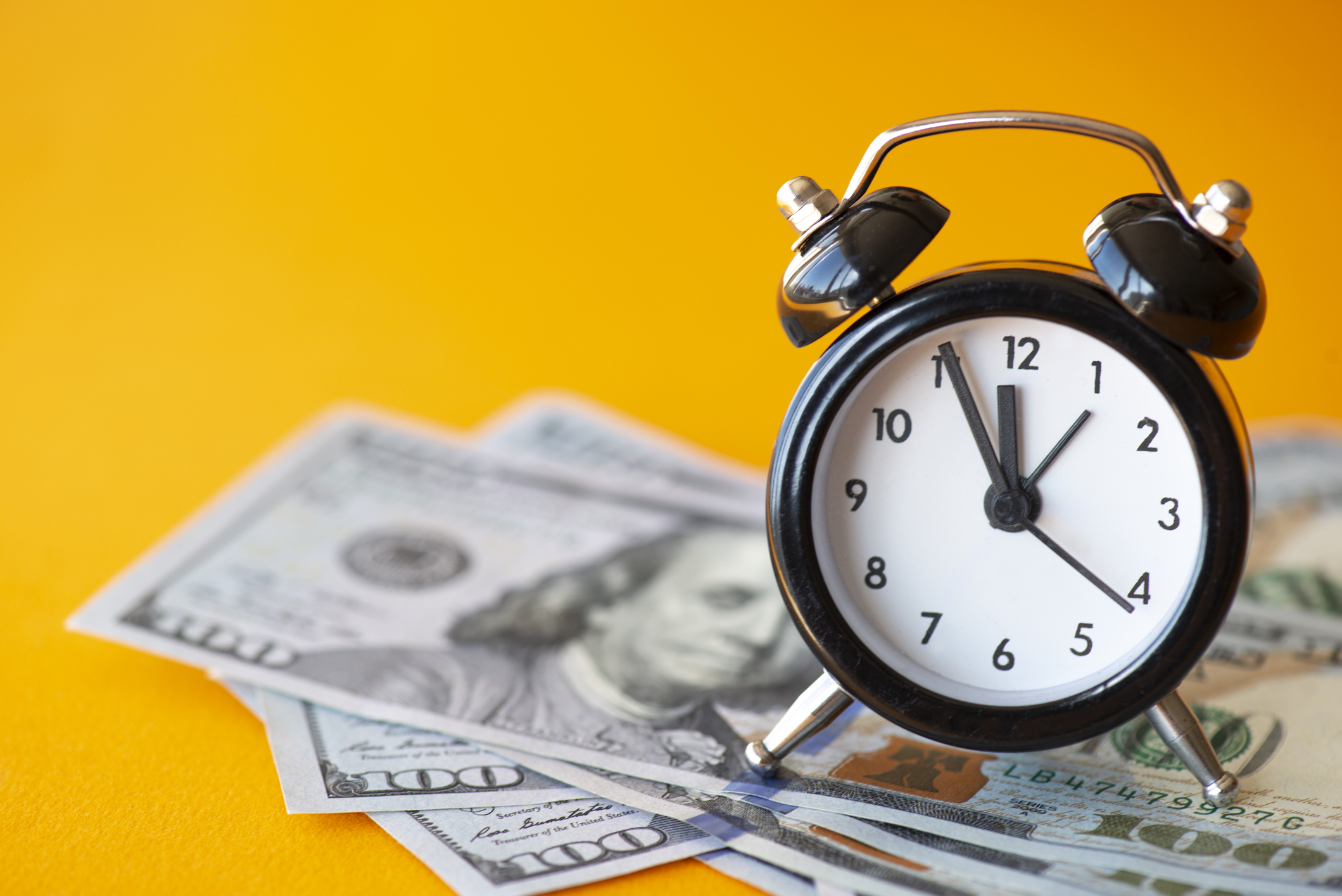 black alarm clock on U.S. bills against a bright yellow background.