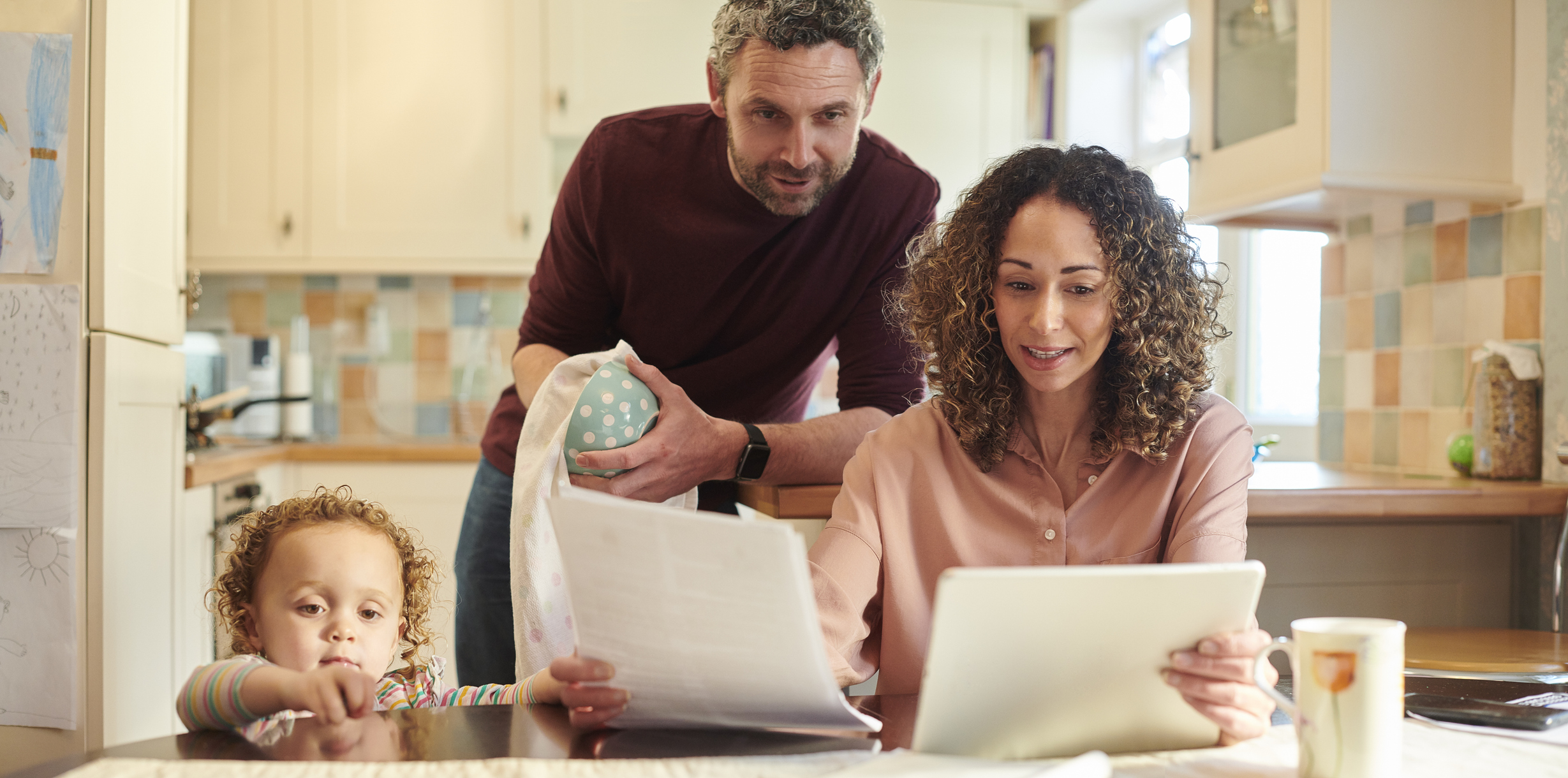 A mother and father look at a computer at the kitchen table while their child looking on.