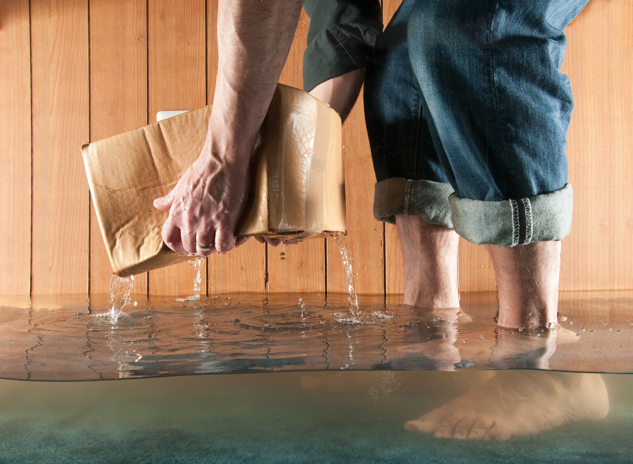Barefoot man in flooded space lifting a wet cardboard box out of the water. 