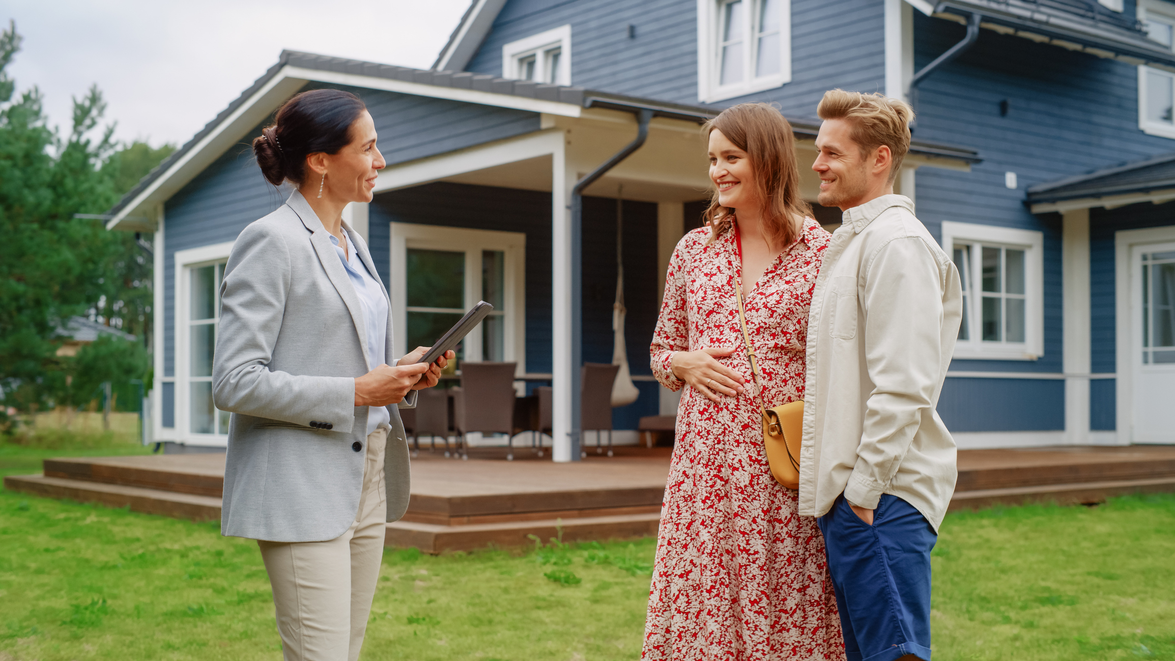 A female real estate agent shows a home to a young couple, exterior view.