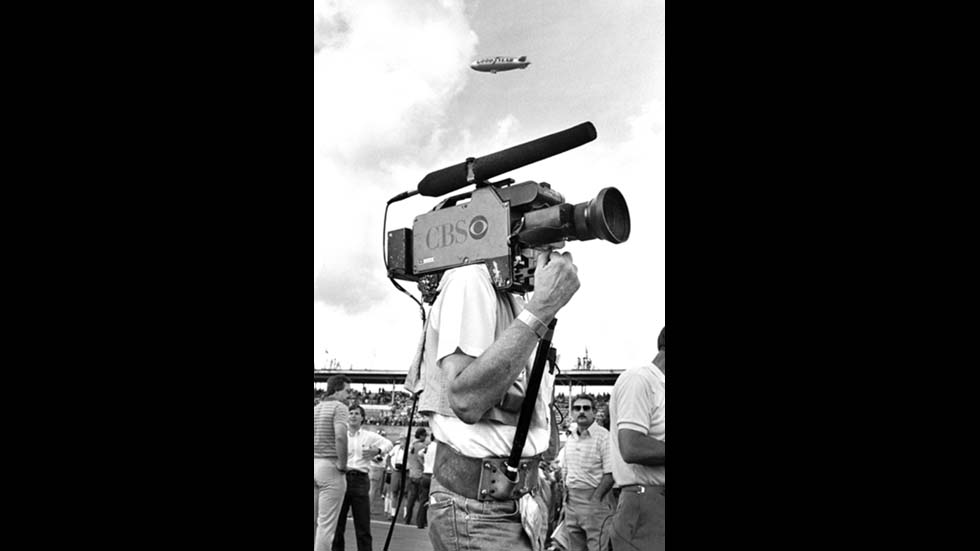 DAYTONA BEACH, FLORIDA - FEBRUARY 1984: A CBS-TV cameraman with an Ikegami video camera on his shoulder prepares to broadcast the 1984 Daytona 500 NASCAR race at Daytona International Speedway in Daytona Beach, Florida. The Goodyear Blimp circles in the air above the speedway. (Photo by Robert Alexander/Getty Images)