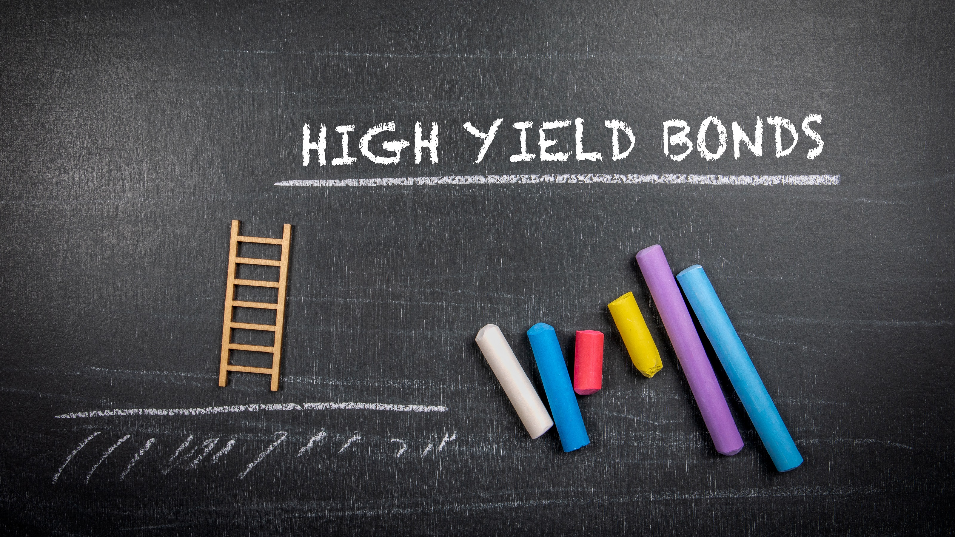 The words high yield bonds on a chalkboard above lined-up pieces of colored chalk.